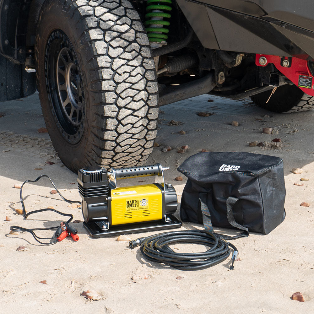  12V air compressor with carry bag and hose setup beside 4WD tyre, ideal for off-road inflation on sandy terrain.