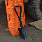 Folding spade leaning against an orange recovery track next to a muddy 4WD tyre.