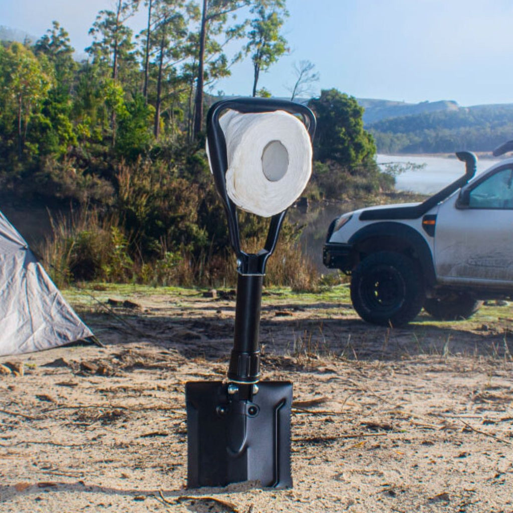 Folding spade in sand holding a toilet paper roll, with a tent and 4WD in the background.