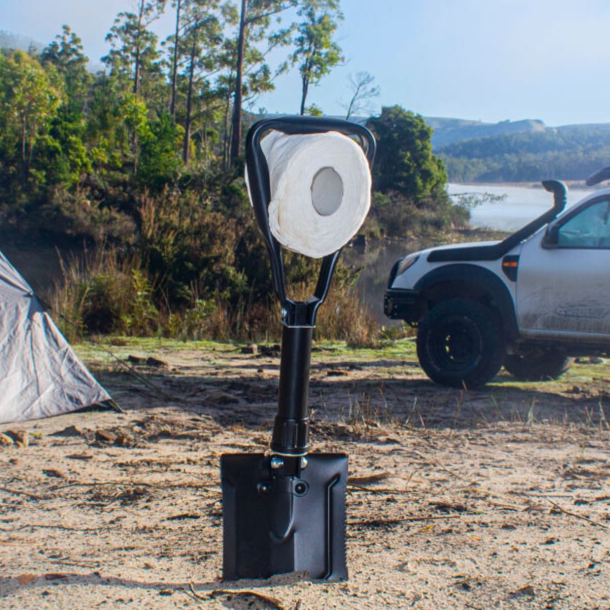 Folding spade in sand holding a toilet paper roll, with a tent and 4WD in the background.