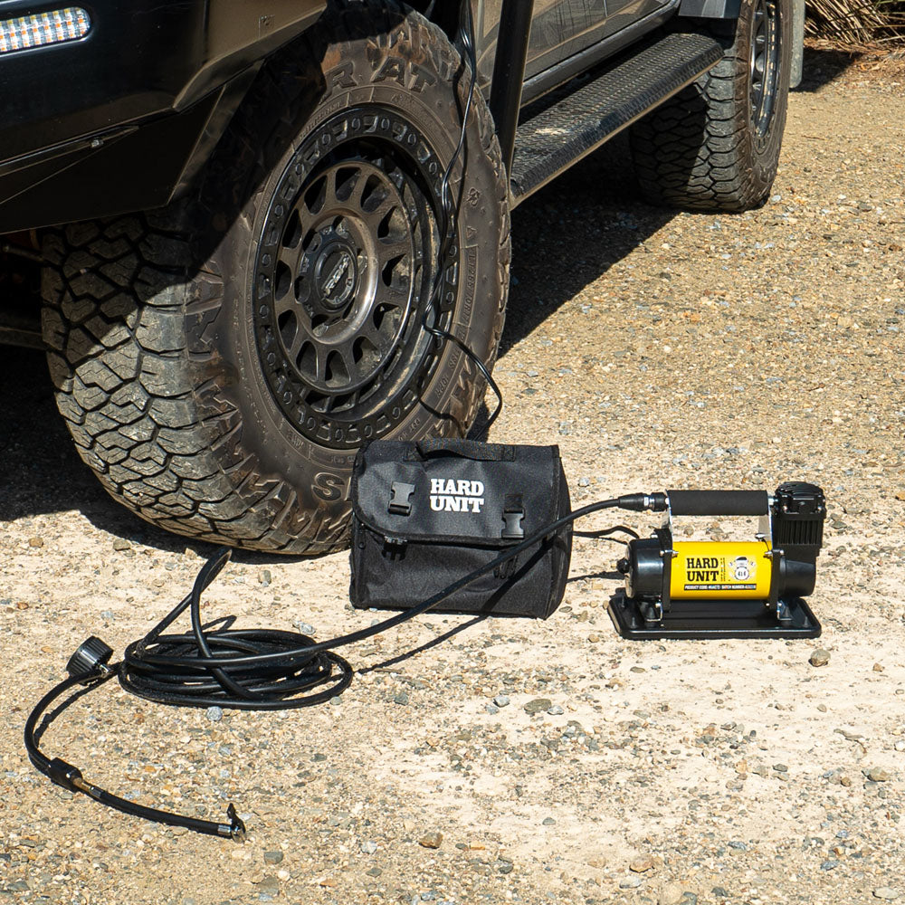 Single 12V air compressor in use inflating a 4WD tyre, shown with carry bag and hose on gravel surface.