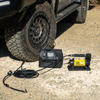 Single 12V air compressor in use inflating a 4WD tyre, shown with carry bag and hose on gravel surface.