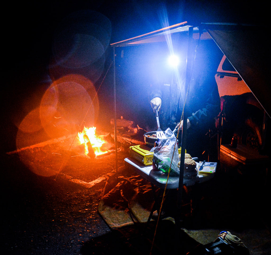 Camper wearing LED beanie headlamp while cooking at night beside campfire under awning, with food prep table and 4WD setup.