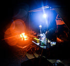 Camper wearing LED beanie headlamp while cooking at night beside campfire under awning, with food prep table and 4WD setup.