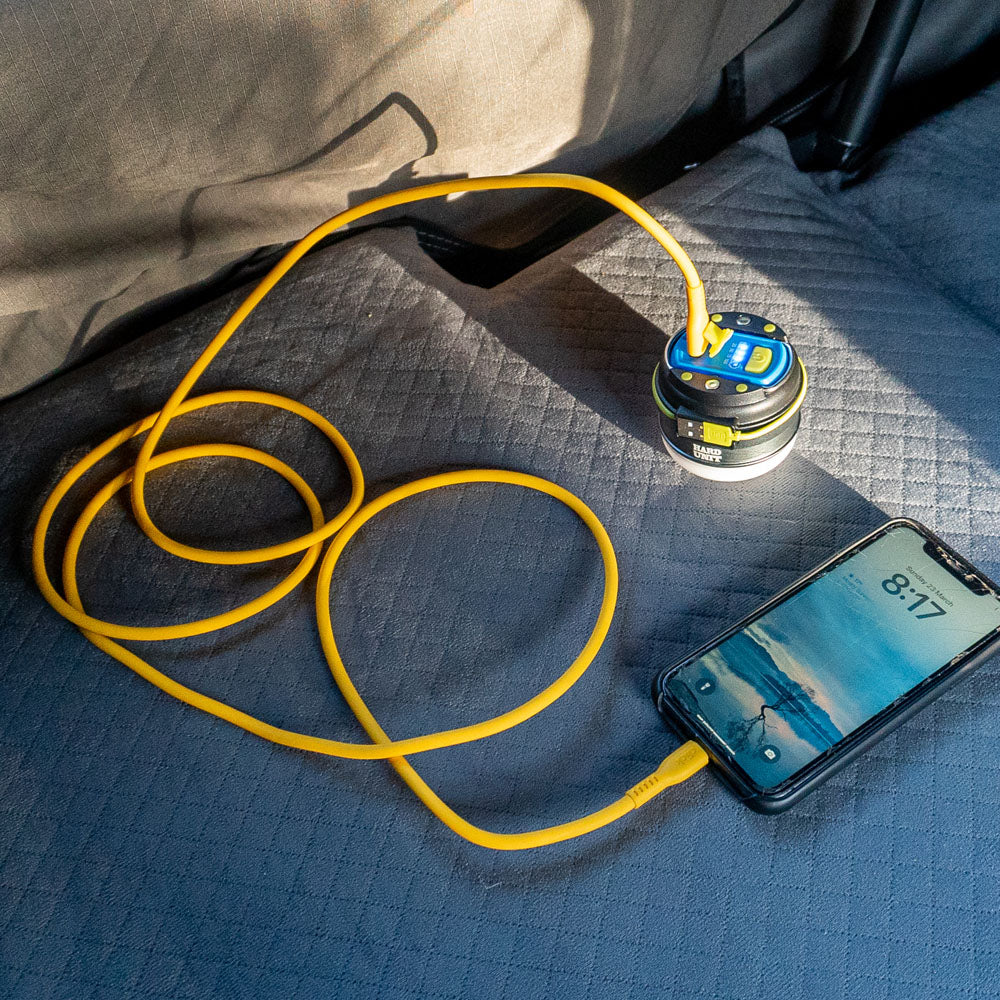 LED camping lantern with USB quick charge function charging a smartphone via yellow cable on quilted car seat