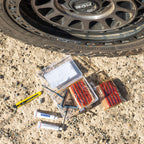 Tubeless tyre repair kit with 56 pieces including repair strings, T-handle tools, rubber cement, utility knife, and plastic storage box next to a 4WD tyre on gravel.