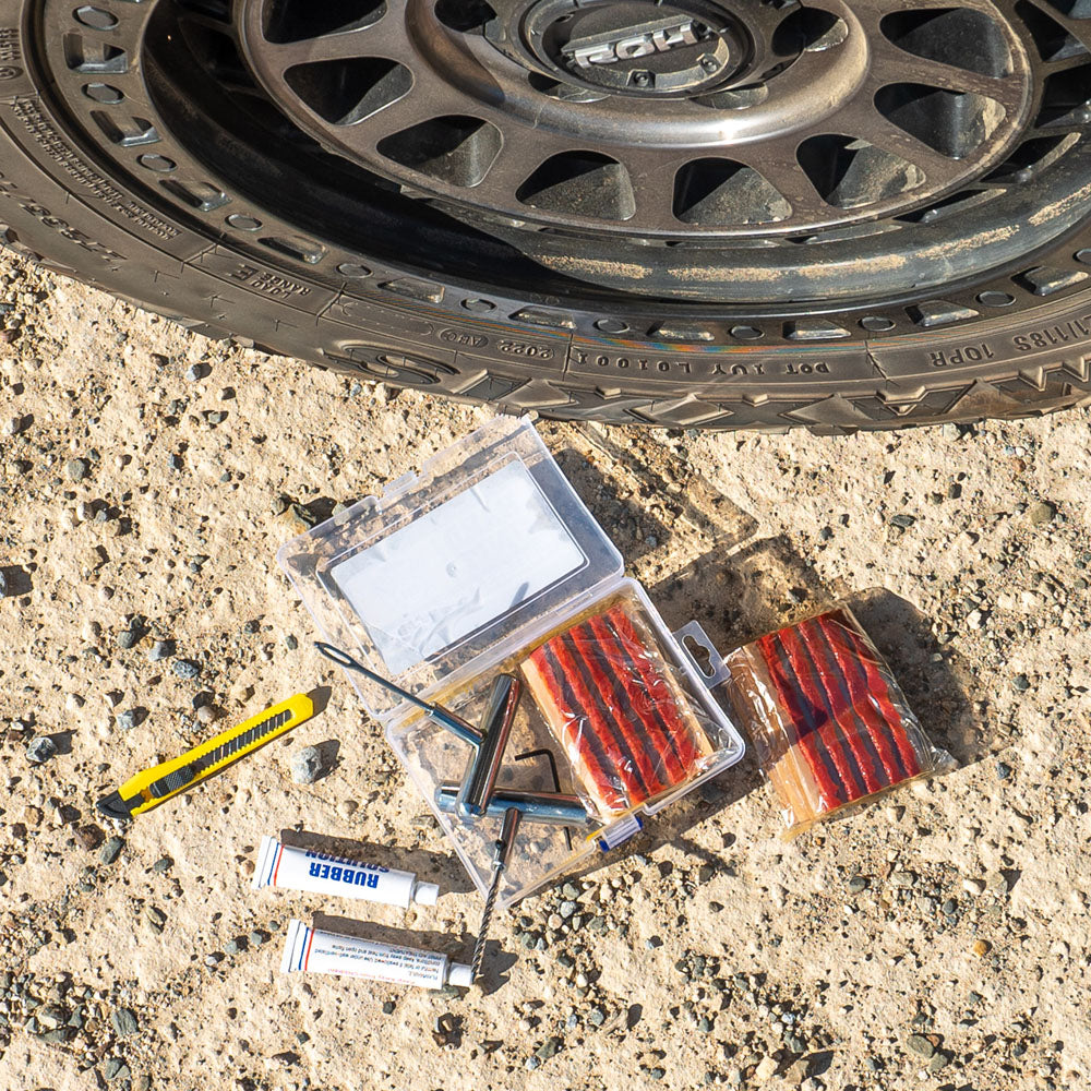 Tubeless tyre repair kit with 56 pieces including repair strings, T-handle tools, rubber cement, utility knife, and plastic storage box next to a 4WD tyre on gravel.