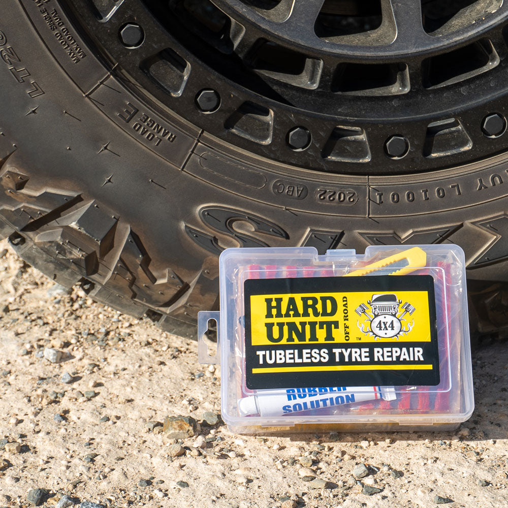Hard Unit branded tubeless tyre repair kit with 56 pieces in a clear plastic case, placed beside a 4WD tyre on gravel.