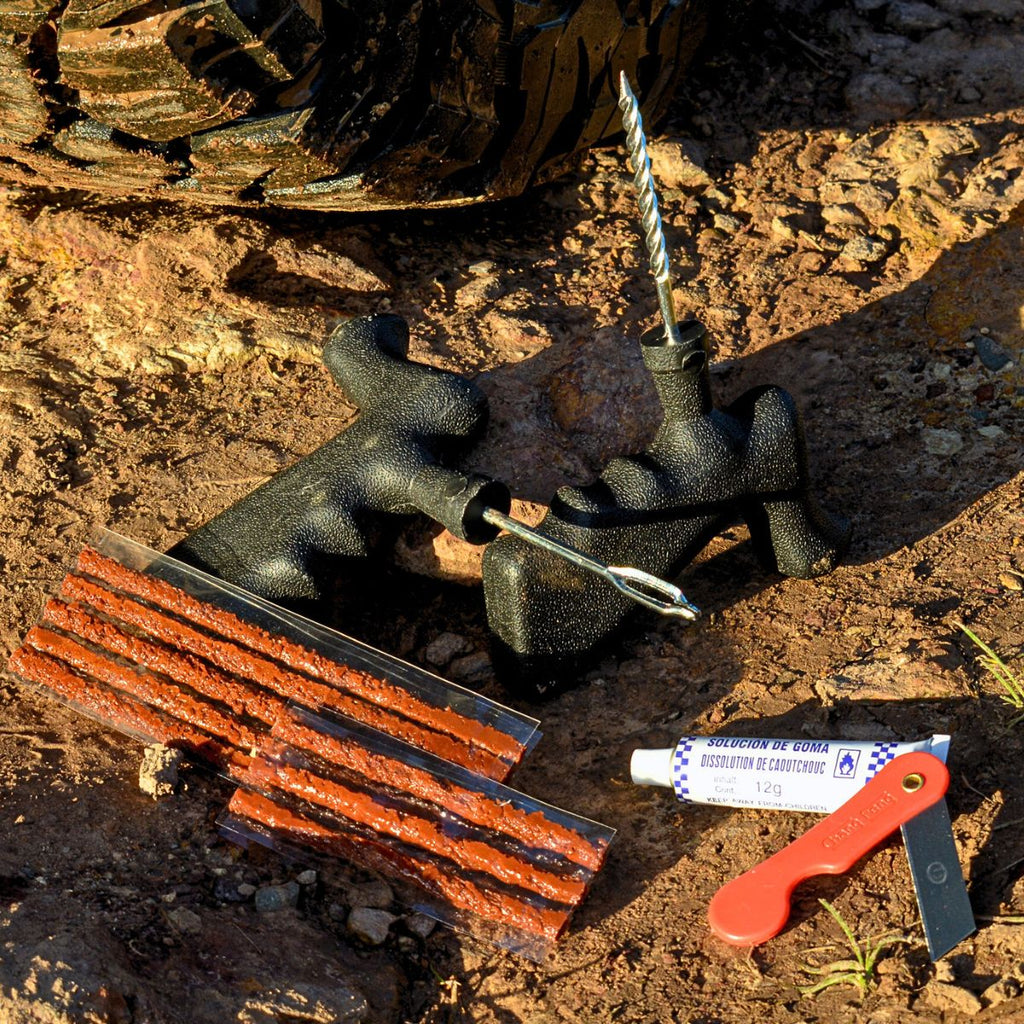 Tyre repair kit laid out on rocky ground, including plug tools, repair strips, rubber solution tube, and a small red blade.