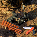 Tyre repair kit laid out on rocky ground, including plug tools, repair strips, rubber solution tube, and a small red blade.