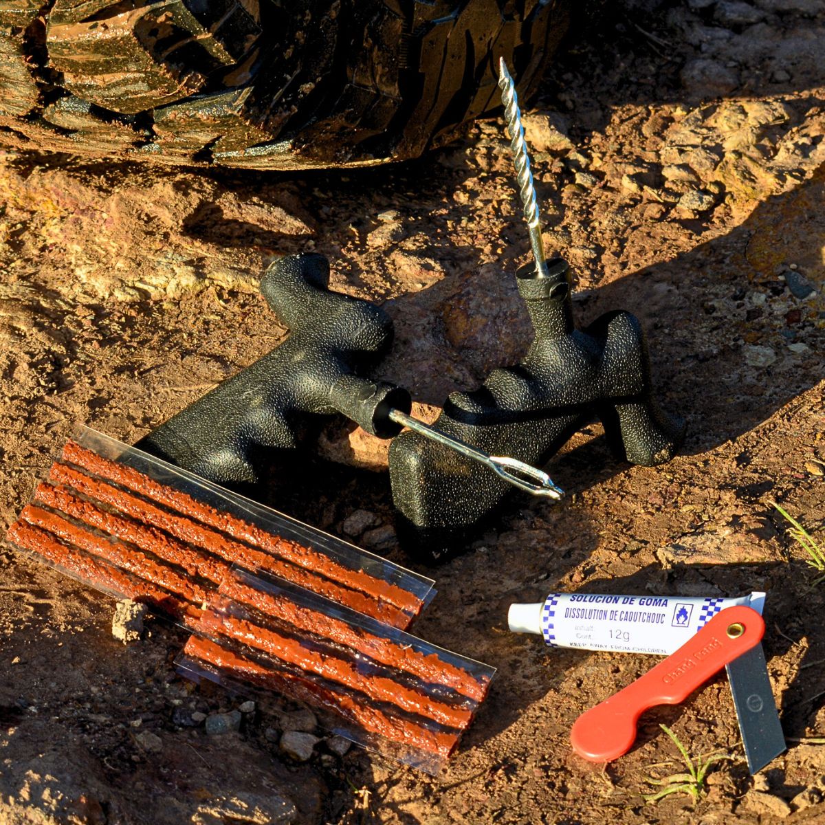 Tyre repair kit laid out on rocky ground, including plug tools, repair strips, rubber solution tube, and a small red blade.