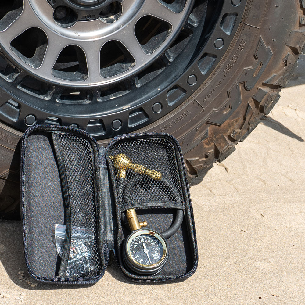 Tyre deflator kit with pressure gauge and accessories in zippered storage case, placed next to a 4WD tyre on sandy terrain.
