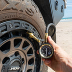 Tyre deflator gauge kit in use on a 4WD tyre at the beach, displaying PSI reading for accurate pressure adjustment.