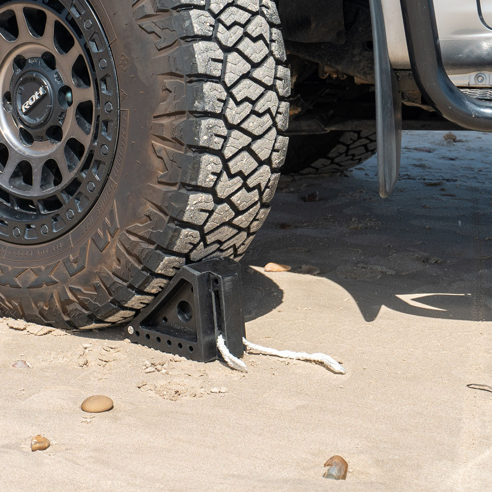 Small solid rubber wheel chock in use under a vehicle tyre on sand, providing stability 