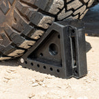 Small solid rubber wheel chock in use under a vehicle tyre on sand, providing stability