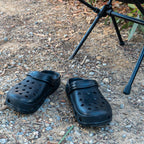 Pair of black off-road clogs with ventilation holes placed on rocky campsite ground beside a folding chair.