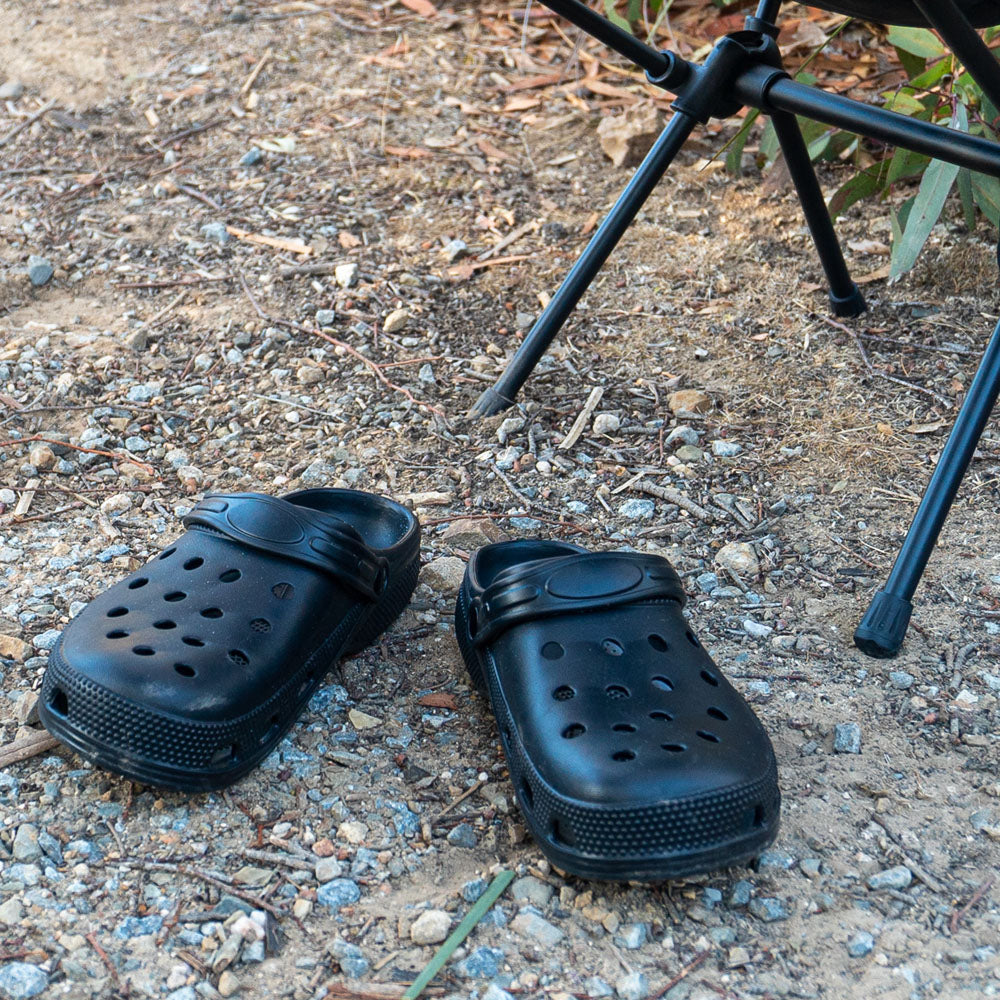 Pair of black off-road clogs with ventilation holes placed on rocky campsite ground beside a folding chair.
